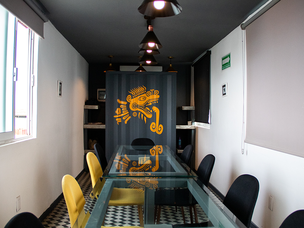 El interior de una moderna sala de juntas construida dentro de un contenedor marítimo, con una larga mesa de vidrio, sillas amarillas y negras, y una pared decorativa con un diseño prehispánico.